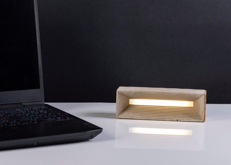 K&ouml;nnte beinhalten: Eine moderne Betonlampe mit rechteckiger Form und einem d&uuml;nnen, wei&szlig;en Lichtstreifen im Inneren. Die Lampe steht auf einer wei&szlig;en Oberfl&auml;che neben einem schwarzen Laptop.