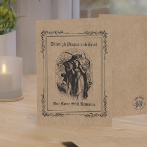 May include: Kraft paper greeting cards with a vintage illustration and text. The front card reads "Through Plague and Trial, Our Love Still Remains" in an ornate frame. A lit candle and eyeglasses are visible.