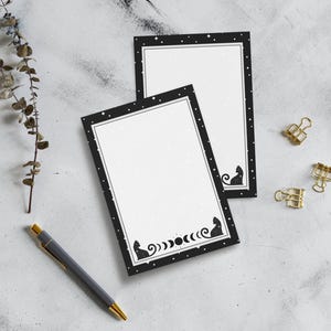 Puede incluir: Dos blocs de notas blancos con bordes negros que presentan un diseño de silueta de gato y fases lunares. Los blocs de notas tienen un patrón de estrellas y están acompañados de un bolígrafo y clips dorados.