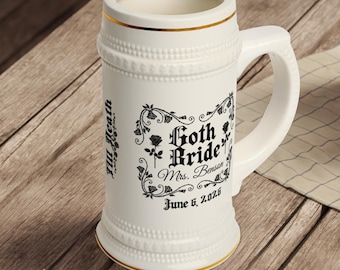 May include: A white ceramic beer stein with a handle, featuring gold trim and embossed detailing. The stein is decorated with the words "Goth Bride" and the name "Mrs. Benson" along with the date "June 6, 2026" in black script.