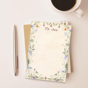 May include: A notepad with a floral border and the text "Ms. Anna" in script. The notepad is surrounded by a silver pen and a cup of coffee. The floral border features blue, orange, and purple flowers.