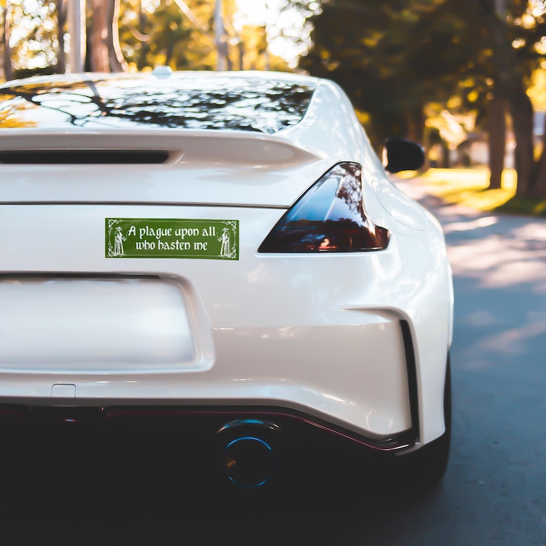 May include: A rectangular green bumper sticker applied to the rear of a white sports car. The sticker features a decorative border, two small figure illustrations on the sides, and the white text: A plague upon all who hasten me.