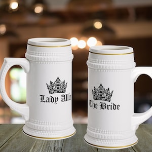 May include: Two white ceramic beer steins with gold rim accents. Each stein features a black crown graphic and text. The left stein displays the text Lady Allie, and the right stein displays the text The Bride. Both steins have a textured base and rim.