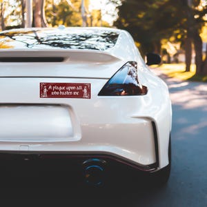 May include: A rectangular maroon bumper sticker applied to the rear of a white sports car. The sticker features a decorative border and white text that reads, A plague upon all who hasten me, flanked by two small illustrations of figures in robes.