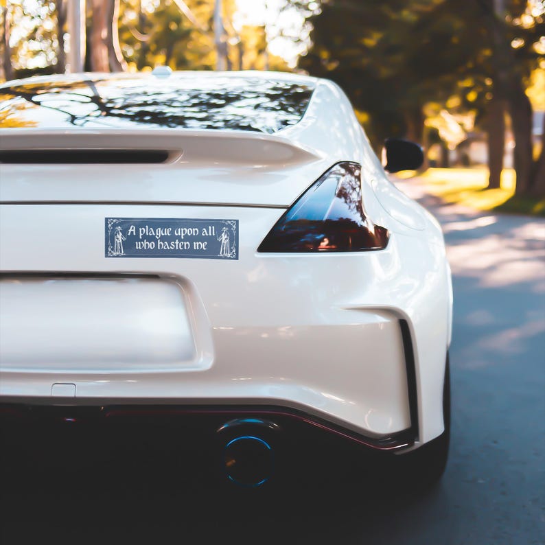 May include: A rectangular bumper sticker applied to the rear of a white sports car. The sticker features a dark blue background with white decorative borders and the text "A plague upon all who hasten me" written in a gothic-style white font.