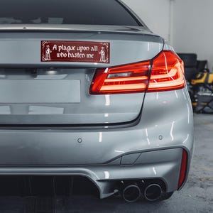 May include: A rectangular bumper sticker with a deep red background and white decorative border. The text reads A plague upon all who hasten me, flanked by two white illustrations of plague doctors in long coats and wide-brimmed hats.