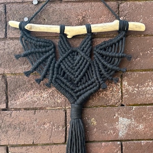 May include: Black macrame wall hanging in the shape of a bat with a wooden branch at the top. The bat has a long tassel hanging from the bottom.