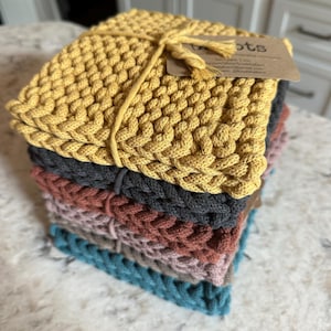 May include: A stack of five knitted pot holders in various colors, including yellow, gray, rust, pink, and teal. The top pot holder is tied with twine and has a small tag that reads "Knots".