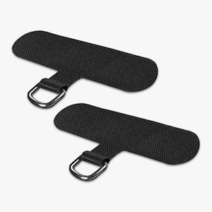 May include: Two black phone lanyard patches with a textured surface and a silver-colored D-ring. The patches are rectangular with rounded ends and a smaller tab with the D-ring attached.