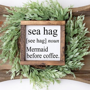 K&ouml;nnte beinhalten: Ein Holzschild mit dem Text "sea hag [see hag] noun Mermaid before coffee." Das Schild liegt auf einem Holztablett mit einem gr&uuml;nen Bl&auml;tterkranz.