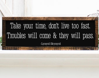 May include: A wooden sign with a black background and white text that reads "Take your time, don't live too fast. Troubles will come & they will pass. -Lynyrd Skynyrd".