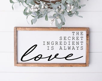 May include: A wooden sign with the text "The Secret Ingredient Is Always Love" in black lettering. The sign is hanging on a white wall with a green and white wreath above it.