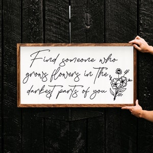 May include: A white wooden sign with a brown frame, featuring the quote "Find someone who grows flowers in the darkest parts of you." with a black floral illustration.