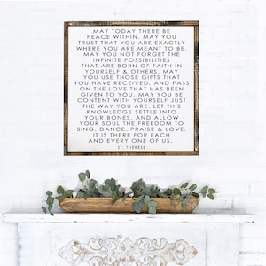 May include: A white wooden sign with a black inspirational quote by Saint Thérèse. The quote reads, "May today there be peace within. May you trust that you are exactly where you are meant to be. May you not forget the infinite possibilities that are born of faith in yourself & others. May you use those gifts that you have received, and pass on the love that has been given to you. May you be content with yourself just the way you are. Let this knowledge settle into your bones, and allow your soul the freedom to sing, dance. Praise & love. It is there for each and every one of us. St. Thérèse"
