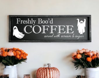 May include: A black framed sign with white text that reads "Freshly Boo'd Coffee served with scream & sugar". Two ghosts are on either side of the text. The sign is hanging on a white wall with fall decorations.