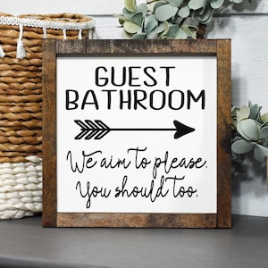 May include: A white wooden sign with a brown frame, featuring the text "GUEST BATHROOM" with a black arrow pointing to the right. The sign also includes the text "We aim to please. You should too."