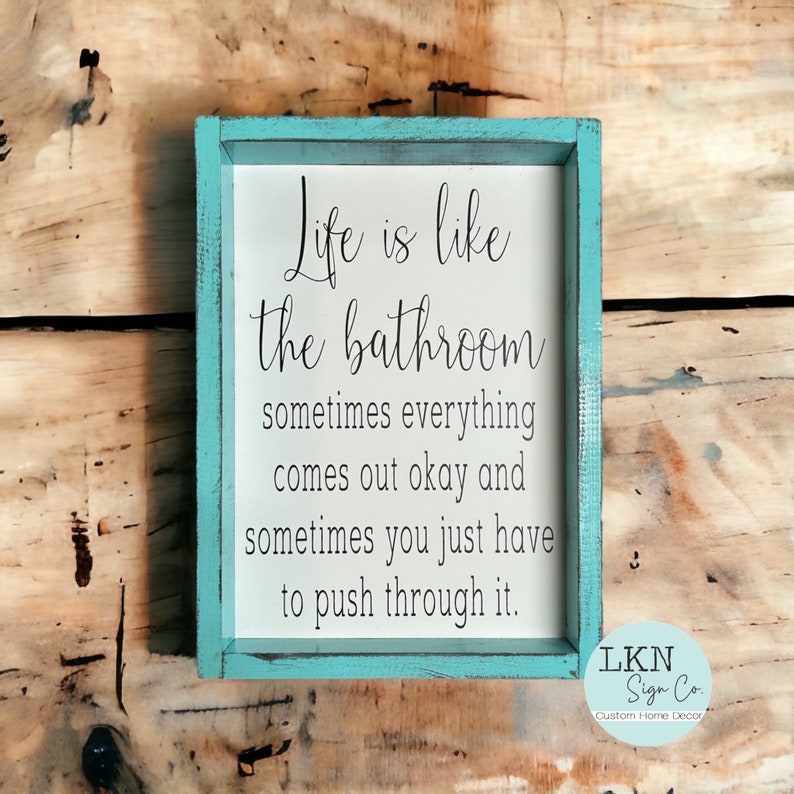 May include: A rectangular wooden sign with a teal frame and white interior. The sign reads "Life is like the bathroom..." in black script. The bottom right corner has the text "LKN Sign Co. Custom Home Decor."