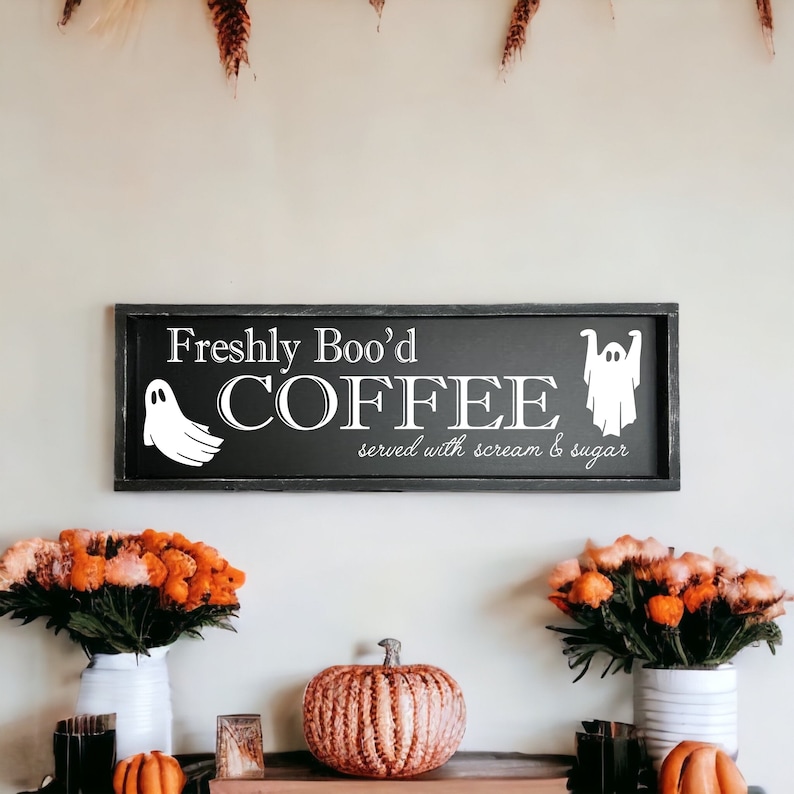 May include: A black framed sign with white text that reads "Freshly Boo'd Coffee served with scream & sugar". Two ghosts are on either side of the text. The sign is hanging on a white wall with fall decorations.