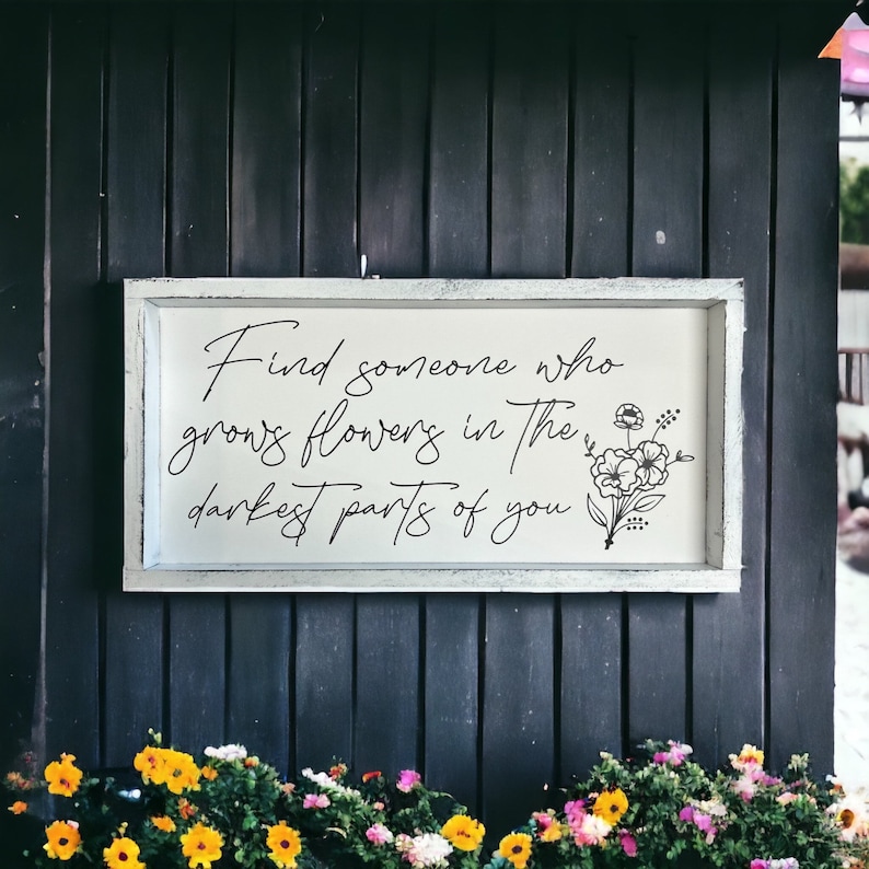 May include: A white wooden sign with black lettering that reads "Find someone who grows flowers in the darkest parts of you". The sign is hanging on a dark gray wooden wall. There are flowers in the foreground.