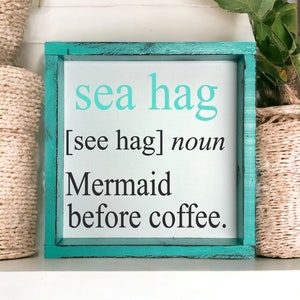 K&ouml;nnte beinhalten: Ein t&uuml;rkisfarbenes Holzschild mit dem Text "sea hag [see hag] noun Mermaid before coffee." Das Schild steht auf einem wei&szlig;en Regal mit Weidenk&ouml;rben auf beiden Seiten.