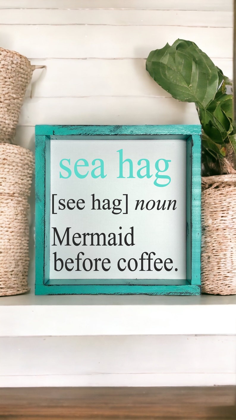 K&ouml;nnte beinhalten: Ein t&uuml;rkisfarbenes Holzschild mit dem Text "sea hag [see hag] noun Mermaid before coffee." Das Schild steht auf einem wei&szlig;en Regal mit Weidenk&ouml;rben auf beiden Seiten.