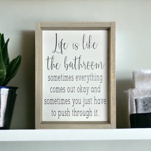 May include: A framed sign with the text "Life is like the bathroom, sometimes everything comes out okay and sometimes you just have to push through it." The frame is light gray, and the background is white.
