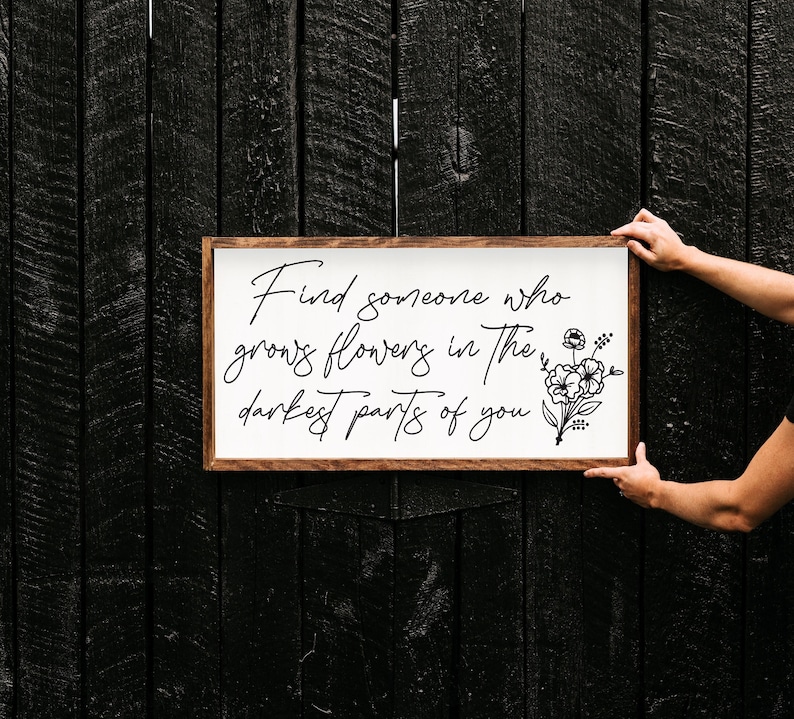 May include: A white wooden sign with a brown frame, featuring the quote "Find someone who grows flowers in the darkest parts of you." with a black floral illustration.