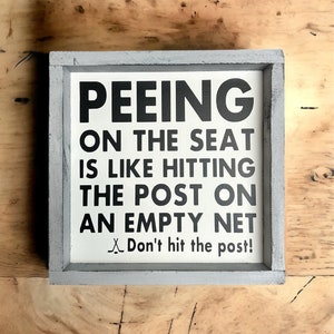 May include: A white wooden sign with black text that reads "Peeing on the seat is like hitting the post on an empty net. Don't hit the post!" The sign is framed in a gray wooden frame.