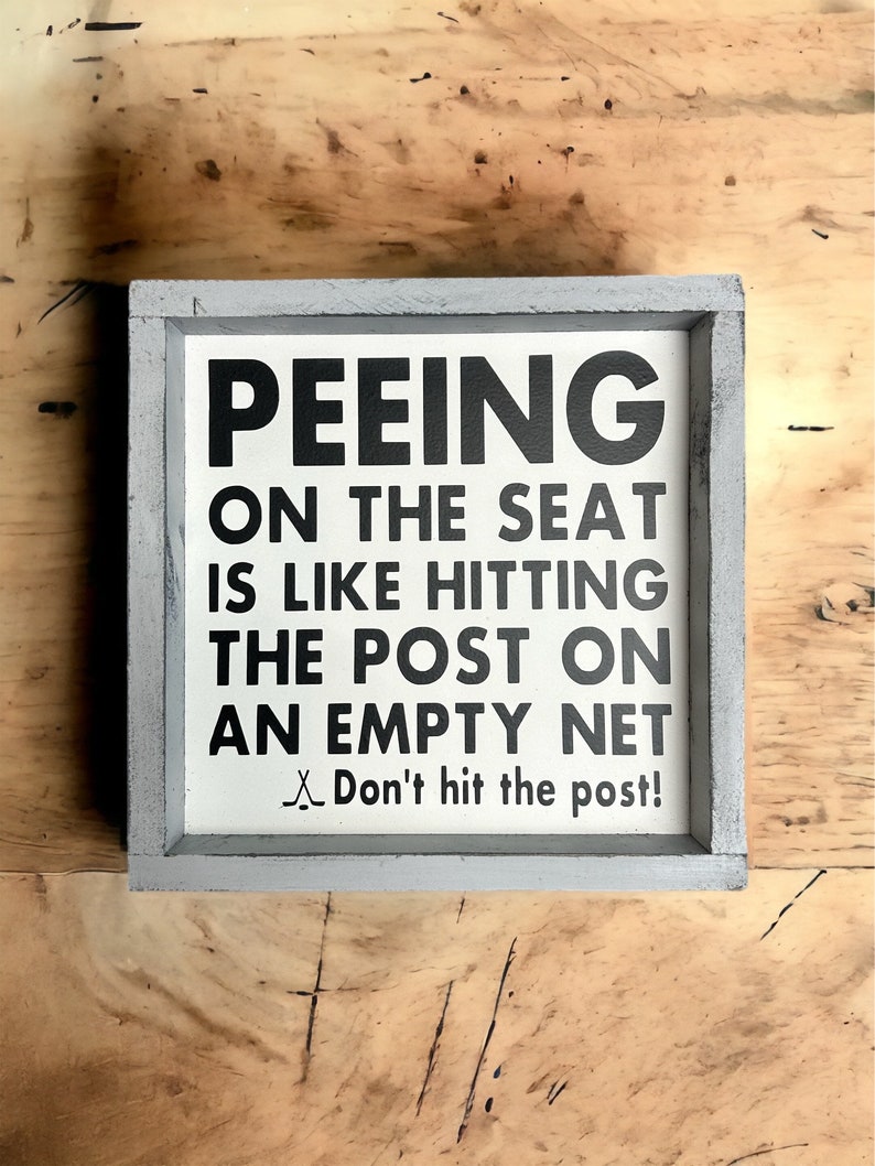 May include: A white wooden sign with black text that reads "Peeing on the seat is like hitting the post on an empty net. Don't hit the post!" The sign is framed in a gray wooden frame.