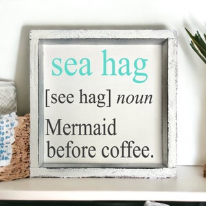 K&ouml;nnte beinhalten: Ein wei&szlig;es Holzschild mit einer abgenutzten Oberfl&auml;che. Das Schild sagt "sea hag [see hag] noun Mermaid before coffee."
