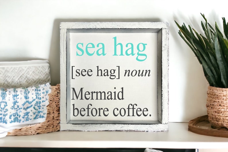 K&ouml;nnte beinhalten: Ein wei&szlig;es Holzschild mit einer abgenutzten Oberfl&auml;che. Das Schild sagt "sea hag [see hag] noun Mermaid before coffee."