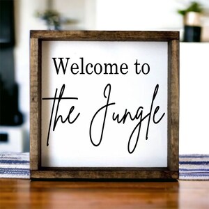 May include: A wooden framed sign with white background and black text that reads "Welcome to the Jungle".