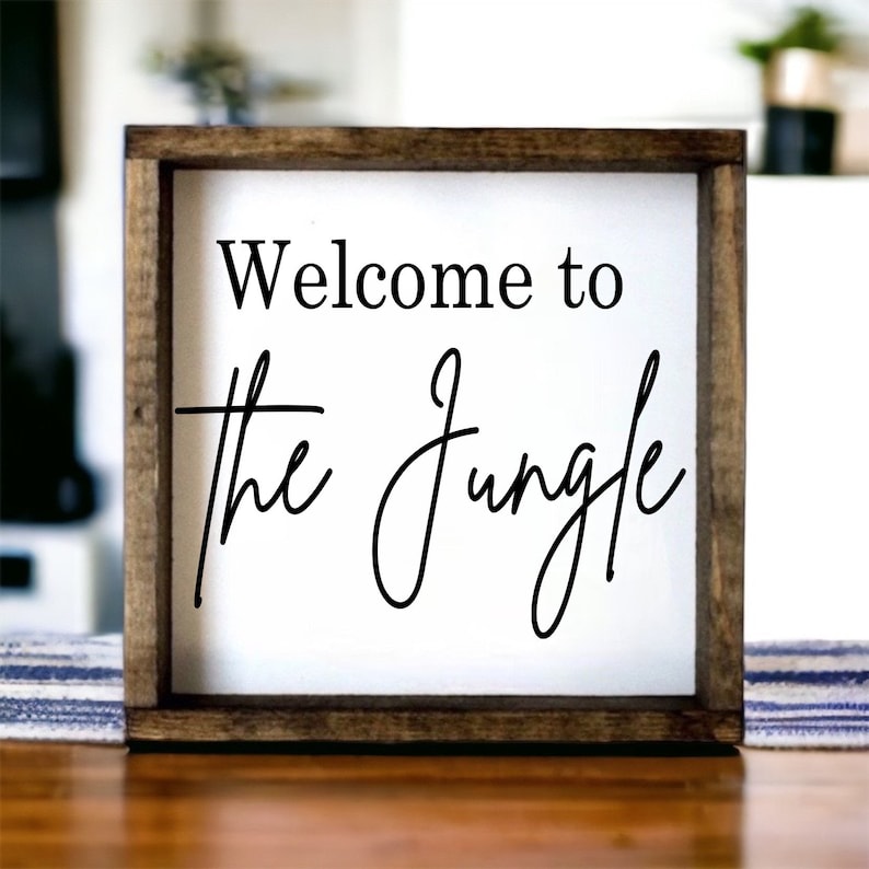 May include: A wooden framed sign with white background and black text that reads "Welcome to the Jungle".