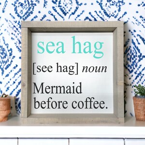 K&ouml;nnte beinhalten: Ein gerahmtes Schild mit dem Text "sea hag [see hag] noun Mermaid before coffee." Das Schild hat einen hellen Holzrahmen und einen wei&szlig;en Hintergrund. Der Text ist in Schwarz und T&uuml;rkis.
