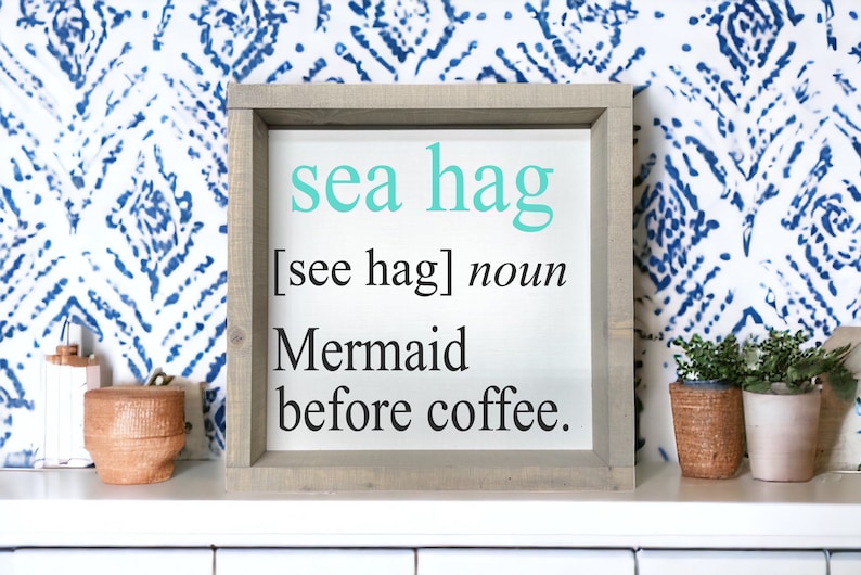 K&ouml;nnte beinhalten: Ein gerahmtes Schild mit dem Text "sea hag [see hag] noun Mermaid before coffee." Das Schild hat einen hellen Holzrahmen und einen wei&szlig;en Hintergrund. Der Text ist in Schwarz und T&uuml;rkis.