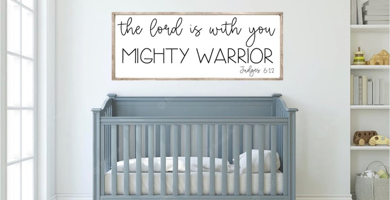 Może przedstawiać: Biały drewniany znak z czarnym tekstem, kt&oacute;ry brzmi "the lord is with you MIGHTY WARRIOR Judges 6:12", wiszący nad szarym ł&oacute;żeczkiem dziecięcym w pokoju dziecięcym.