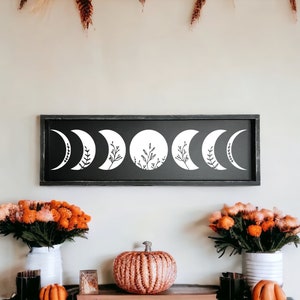 May include: A black rectangular wooden sign displaying a white moon phases design with botanical details. The phases are arranged horizontally. The sign is displayed above a decorative arrangement with flowers and pumpkins.