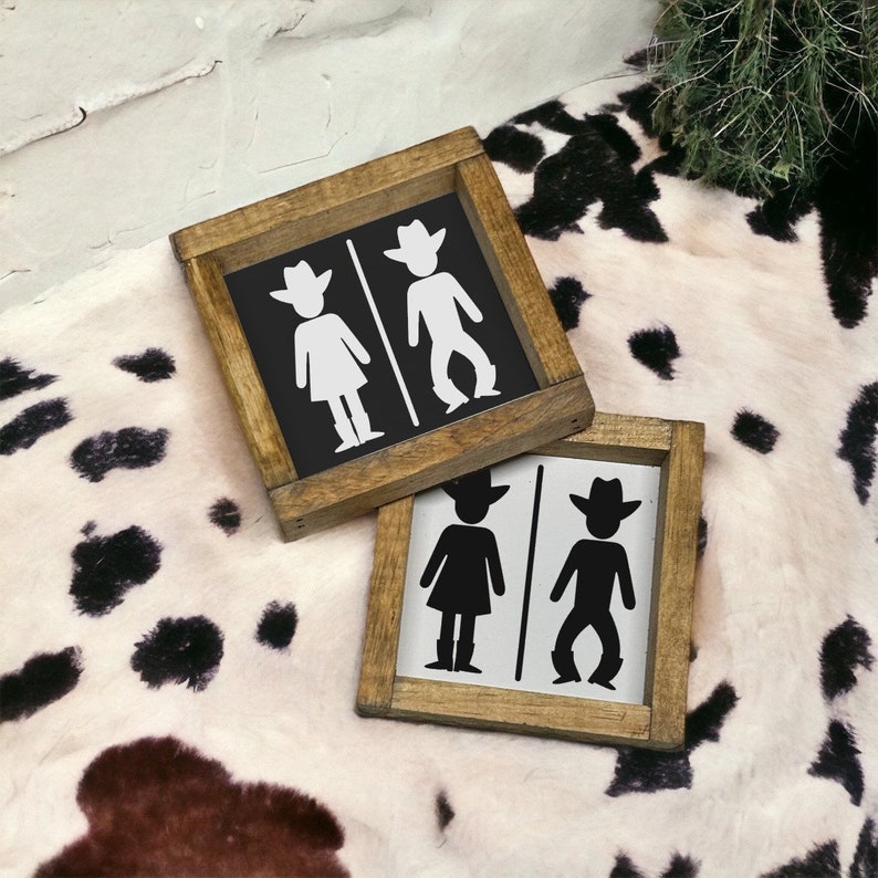 Op de afbeelding: Twee houten borden met een zwarte achtergrond en witte cowboy-silhouetten. De borden zijn gelabeld met "Dames" en "Heren" met een cowboyhoed op elk figuur. De borden liggen op een wit en zwart koeienhuidkleed.