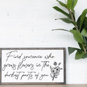 May include: A framed sign with a white background and black text that reads "Find someone who grows flowers in the darkest parts of you". The sign is framed in a dark wood frame and has a black and white floral illustration in the bottom right corner.