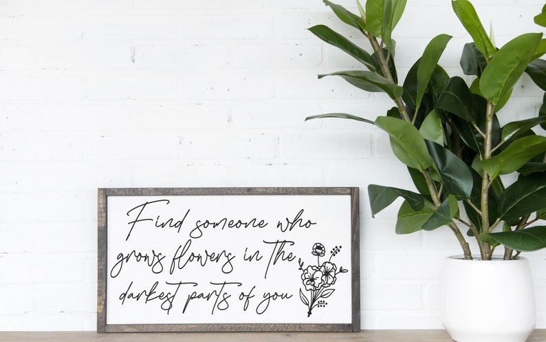 May include: A framed sign with a white background and black text that reads "Find someone who grows flowers in the darkest parts of you". The sign is framed in a dark wood frame and has a black and white floral illustration in the bottom right corner.