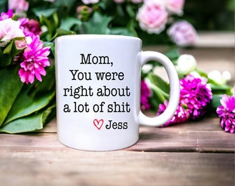May include: White ceramic coffee mug with black text that reads "Mom, You were right about a lot of shit ♥ Jess". The mug is sitting on a wooden surface with pink flowers in the background.