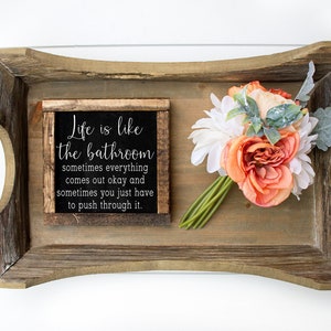 May include: A rustic wooden tray with handles made of wooden beads. The tray holds a small framed sign with the text "Life is like the bathroom" and a bouquet of orange and white flowers.