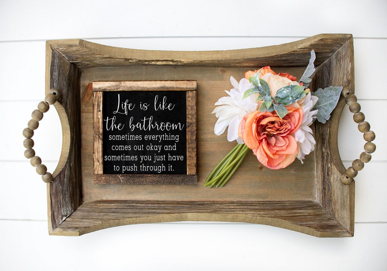 May include: A rustic wooden tray with handles made of wooden beads. The tray holds a small framed sign with the text "Life is like the bathroom" and a bouquet of orange and white flowers.