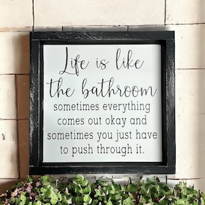 May include: A framed sign with the text "Life is like the bathroom, sometimes everything comes out okay and sometimes you just have to push through it." The sign is black and white and is above three small potted plants in a woven basket.