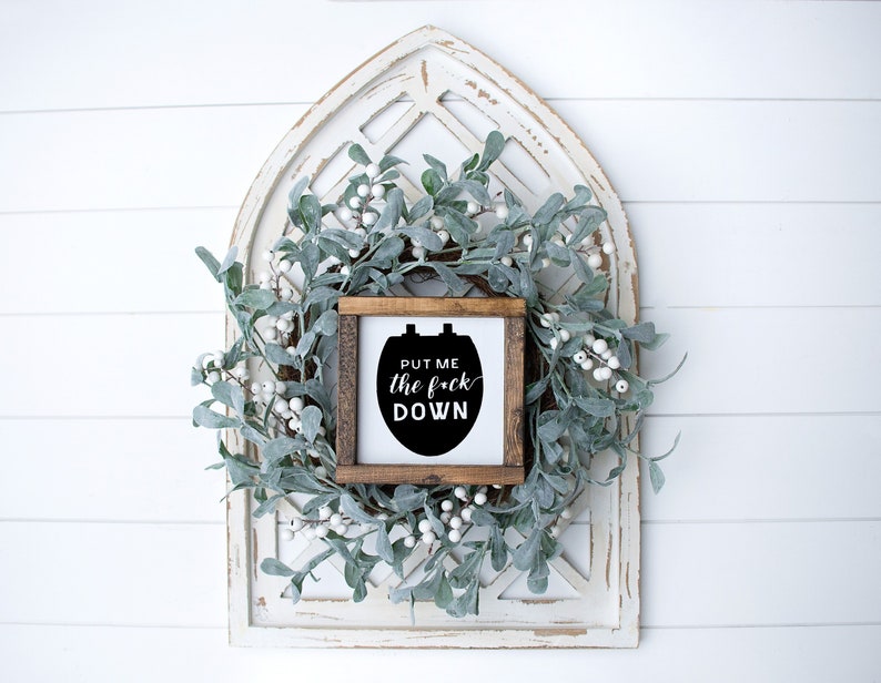 May include: A wooden sign with a black silhouette of a toilet seat and the text "Put me the f*ck down". The sign is hanging on a white wall with a white and green wreath.