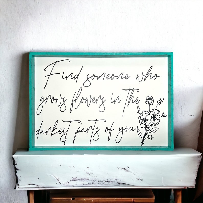 May include: A teal framed sign with the text "Find someone who grows flowers in the darkest parts of you."  The text is in black cursive font and there is a black floral design in the bottom right corner.