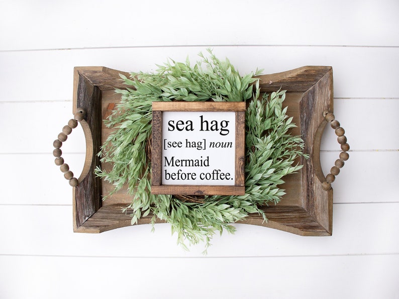 K&ouml;nnte beinhalten: Ein Holzschild mit dem Text "sea hag [see hag] noun Mermaid before coffee." Das Schild liegt auf einem Holztablett mit einem gr&uuml;nen Bl&auml;tterkranz.