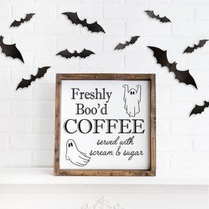 K&ouml;nnte beinhalten: Ein Holzschild mit wei&szlig;em Hintergrund und schwarzem Text, der "Freshly Boo'd COFFEE served with scream & sugar" lautet. Das Schild ist mit zwei Geisterillustrationen verziert und von schwarzen Flederm&auml;usen an einer wei&szlig;en Backsteinwand umgeben.