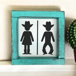 Op de afbeelding: Een turquoise houten bord met een witte achtergrond en zwarte silhouetten van een cowboy en een cowgirl die cowboyhoeden en laarzen dragen. Het bord zegt "Dames" en "Heren" met een pijl die naar elk silhouet wijst.