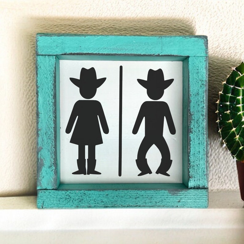 Op de afbeelding: Een turquoise houten bord met een witte achtergrond en zwarte silhouetten van een cowboy en een cowgirl die cowboyhoeden en laarzen dragen. Het bord zegt "Dames" en "Heren" met een pijl die naar elk silhouet wijst.